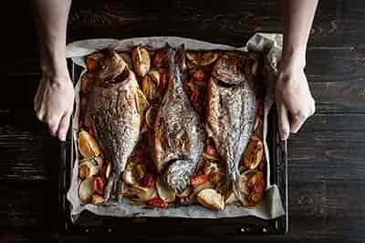 3 full body fish with potatoes onions spices on serving pan coming out of oven ready to eat